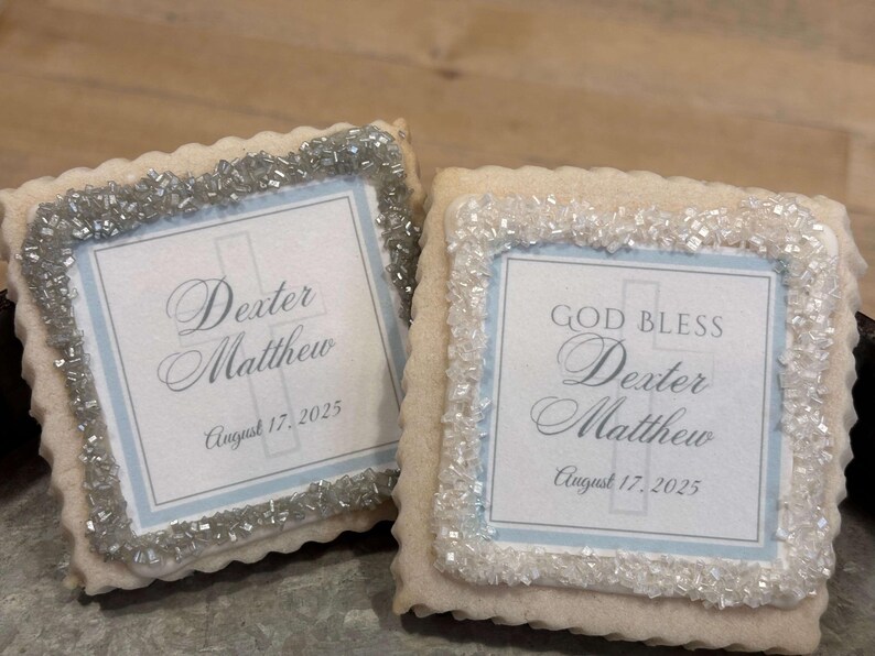 May include: Two decorated sugar cookies with white frosting and silver sprinkles. The cookies have a blue and white design with a cross and the text "God Bless Dexter Matthew August 17, 2025".