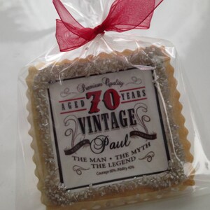 K&ouml;nnte beinhalten: Ein quadratischer Zuckerkeks, der mit silbernen Streuseln und einem Druckdesign verziert ist, auf dem "Premium Quality Aged 70 Years Vintage Paul The Man - The Myth The Legend Courage 60% / Ability 40%" steht.