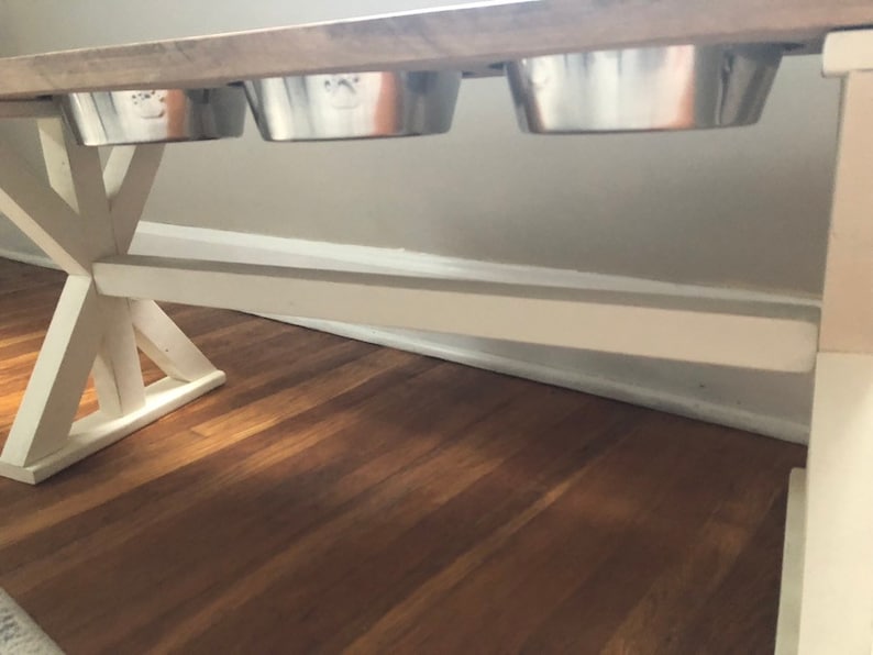 May include: White wooden table with a distressed wood top. Three stainless steel dog bowls are attached to the underside of the table.