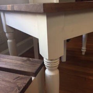 May include: A close-up of a white and brown wooden table with a rectangular top. The table has four white, turned legs. The tabletop is a dark brown color. The table is near a window and a wooden bench.