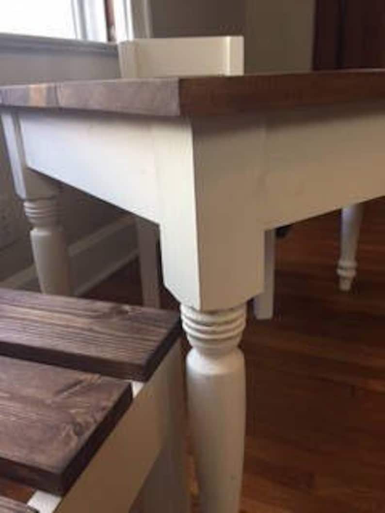 May include: A close-up of a white and brown wooden table with a rectangular top. The table has four white, turned legs. The tabletop is a dark brown color. The table is near a window and a wooden bench.