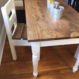 May include: A small, white-legged wooden table with a matching chair attached. The tabletop is stained a medium brown and has three small white containers with colorful markers and pens. The table is on a hardwood floor.