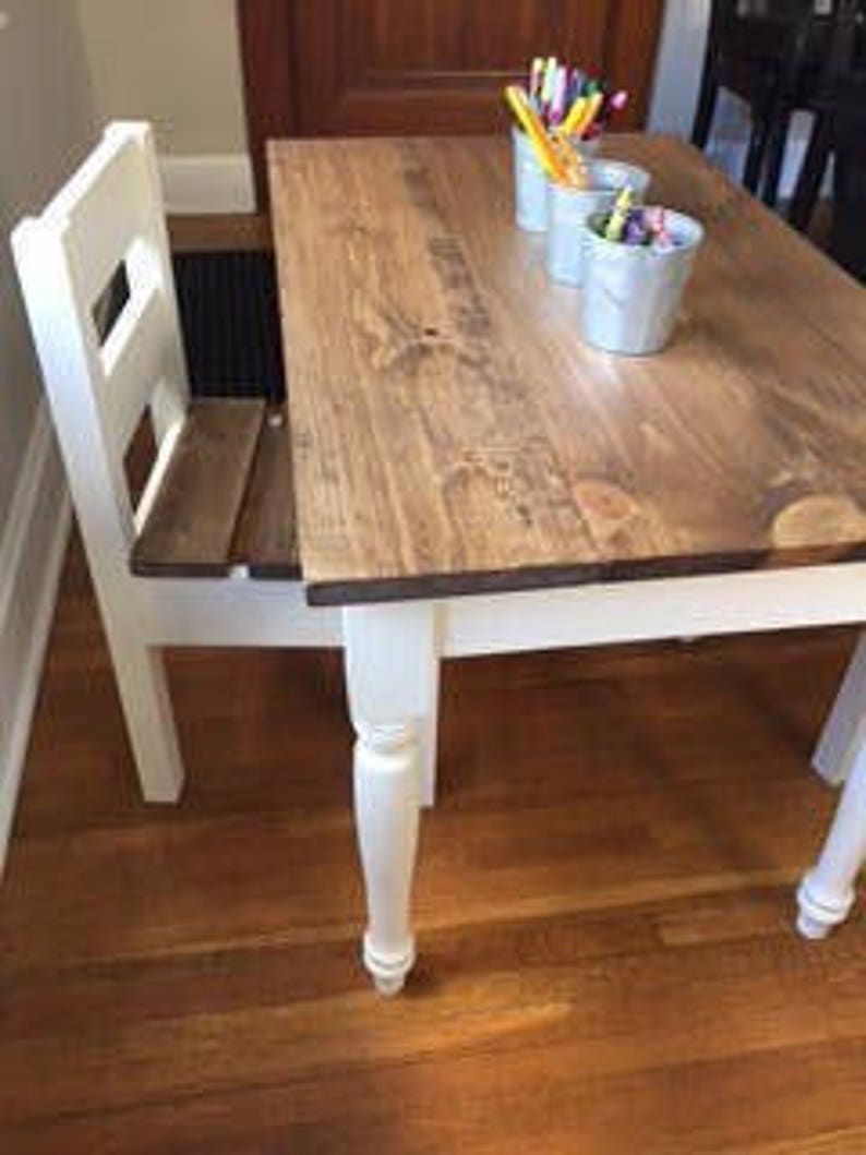 May include: A small, white-legged wooden table with a matching chair attached. The tabletop is stained a medium brown and has three small white containers with colorful markers and pens. The table is on a hardwood floor.