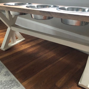 May include: A white wooden dog food stand with three stainless steel bowls. The stand has an X-shaped base and a wooden top.