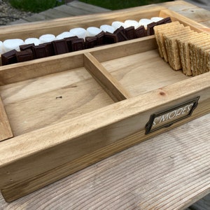 May include: A wooden tray with three compartments for making s'mores. The tray has a metal label that says "S'mores". The compartments are filled with graham crackers, chocolate squares, and marshmallows.
