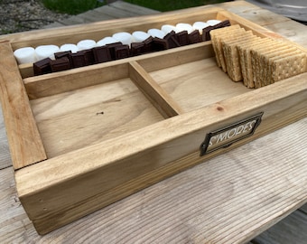 May include: A wooden tray with three compartments for making s'mores. The tray has a metal label that says "S'mores". The compartments are filled with graham crackers, chocolate squares, and marshmallows.