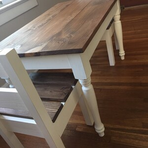 May include: A small wooden table with a dark brown tabletop and white legs. The table has a classic design, with turned legs and a simple, functional style. The table is approximately 30 inches long and 20 inches wide.