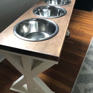 May include: A wooden dog food stand with three stainless steel bowls. The stand has a white X-shaped base and a brown top.