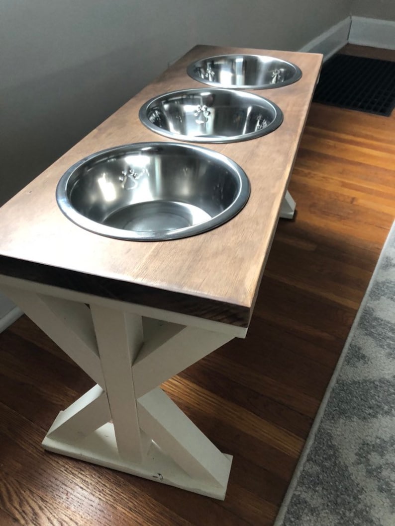 May include: A wooden dog food stand with three stainless steel bowls. The stand has a white X-shaped base and a brown top.