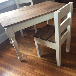 May include: A small wooden table with a light brown tabletop and white legs. A matching chair with a dark brown seat and white frame is positioned at the table. The table is approximately 36 inches long.