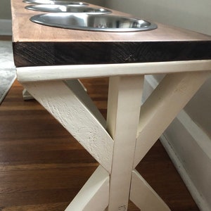 May include: A wooden dog bowl stand with three stainless steel bowls. The stand has a white painted base with an X-shaped design. The top of the stand is a natural wood color.