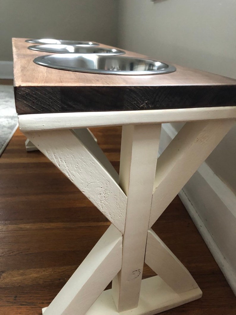 May include: A wooden dog bowl stand with three stainless steel bowls. The stand has a white painted base with an X-shaped design. The top of the stand is a natural wood color.