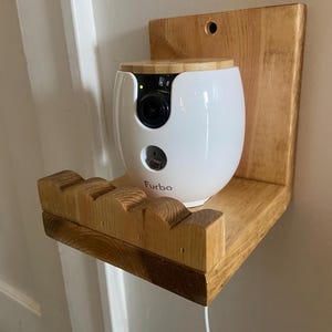 May include: A white Furbo pet camera with a bamboo lid sits on a light brown wooden wall-mounted shelf. The shelf features a scalloped front edge and a vertical back panel with a pre-drilled hole for mounting. A white power cord hangs downward.