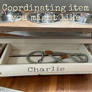 May include: White wooden dog food stand with three stainless steel bowls and a pull-out drawer labeled "Charlie". A green and brown dog leash is in the drawer.