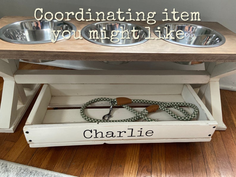 May include: White wooden dog food stand with three stainless steel bowls and a pull-out drawer labeled "Charlie". A green and brown dog leash is in the drawer.