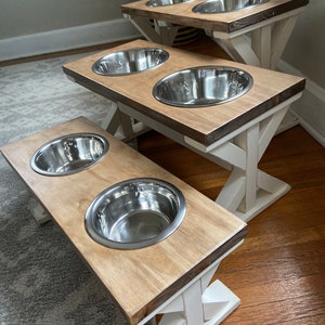 May include: Three elevated dog bowls with white painted legs and a natural wood top. The bowls are made of stainless steel and are arranged in a staggered formation.