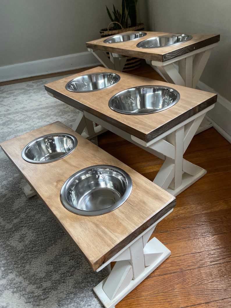 May include: Three elevated dog bowls with white painted legs and a natural wood top. The bowls are made of stainless steel and are arranged in a staggered formation.