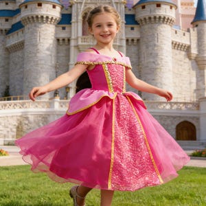 May include: A young girl in a pink princess dress with gold trim and a sparkling bodice, twirling on a green lawn. The dress has a layered skirt and off-the-shoulder sleeves. A fairytale castle is in the background.