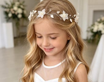 May include: A delicate floral headband featuring white fabric flowers, small pearls, and clear crystals. The headband is on a thin silver wire and is worn by a person with long, wavy blonde hair. The person is wearing a white dress.