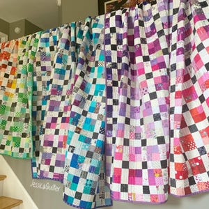May include: Five colorful patchwork quilts with a checkerboard pattern in black and white. The quilts are hanging on a railing and are made of various fabrics in shades of green, blue, purple, and pink.