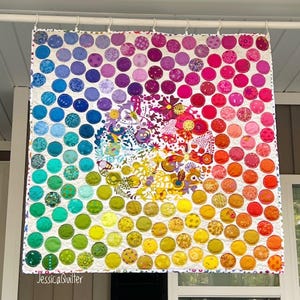 Könnte beinhalten: Ein farbenfroher Quilt mit einem kreisförmigen Muster aus Stoffkreisen in Blautönen, Grün, Gelb, Orange, Rot und Pink. Die Kreise sind spiralförmig angeordnet, wobei sich in der Mitte der Spirale ein weißer Kreis mit einem Blumendesign befindet. Der Quilt hängt an einer weißen Stange.