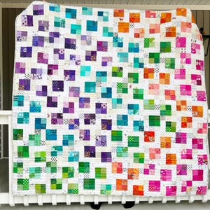 May include: A colorful patchwork quilt with a rainbow gradient of squares. The quilt is made of various fabrics in shades of purple, blue, green, orange, and pink. The quilt is hanging on a white porch railing.