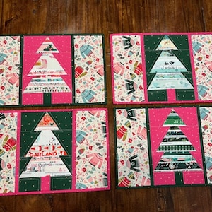 Op de afbeelding: Vier kerstboom placemats. Elk heeft een patchwork boomontwerp in groen en wit op een roze achtergrond. De zijkanten hebben een patroon van truien en andere vakantieartikelen. De placemats liggen op een houten oppervlak.