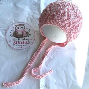 Op de afbeelding: Een lichtroze gebreide babymuts met een ingewikkeld kabelpatroon en twee lange bijpassende gebreide linten. Naast de muts ligt een ronde witte sticker met een illustratie van een uil en de tekst The Nest of Stitches.