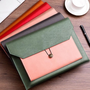 Könnte beinhalten: Ein Stapel bunter Dokumentenmappen in den Farben Orange, Rot, Rosa, Grau und Grün. Die oberste Mappe ist grün mit rosa Innenraum und Knopfverschluss. Ein Laptop, ein Stift und eine Teetasse sind ebenfalls zu sehen.