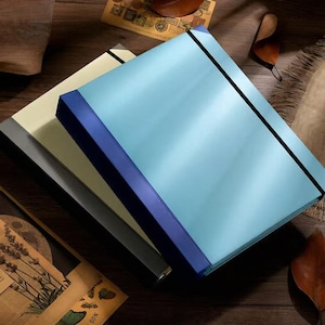 May include: A stack of three rectangular document folders featuring a smooth, solid-colored finish. The top folder is light blue, the middle folder is cream, and the bottom folder is dark gray. Each folder is secured with a thin black elastic band.