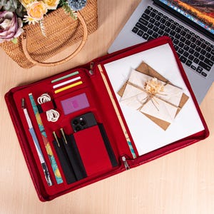 May include: A red leather portfolio with multiple compartments, including card slots, pen holders, and a phone pocket. The portfolio is open, revealing a laptop, stationery, and a woven handbag with flowers.