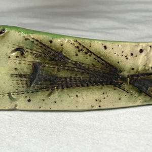 Puede incluir: Un plato de cerámica hecho a mano con forma rectangular y borde verde claro. El centro presenta un diseño texturizado con marcas marrón oscuro y negro, y pequeñas motas negras. El plato tiene una apariencia artística única.