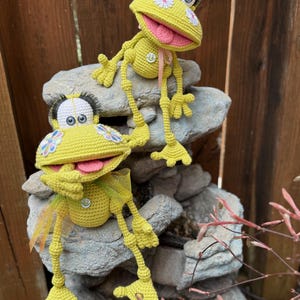 May include: Two crocheted yellow frog plushies with pink tongues and white eyes, sitting on a pile of gray rocks. The frogs have flowers and smiley faces on their bodies.