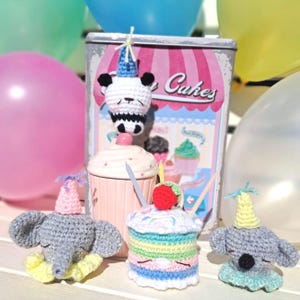 Könnte beinhalten: Eine Sammlung von drei gehäkelten Tierfiguren, darunter ein Panda, ein Elefant und ein Koala, die jeweils einen Partyhut tragen. Ein gehäkelter Cupcake mit Erdbeer-Topping und zwei Kerzen steht in der Mitte vor einer Metalldose.