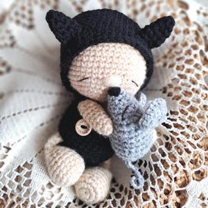 May include: A handmade crochet doll wearing a black hat with ears and a black outfit, hugging a gray crochet mouse. The doll has a beige face, arms, and legs. A small wooden button with text is on the doll's outfit.