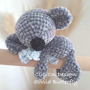 Könnte beinhalten: Ein graues gehäkeltes Koala-Plüschtier mit schwarzer Nase und geschlossenen Augen, das über ein Clip-Design verfügt. Der Text Clip-On Design @Wild Butterfly ist in Weiß unten auf dem Koala aufgedruckt, der auf einem Holzstab ruht.