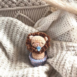 Könnte beinhalten: Ein handgefertigtes gehäkeltes Löwenspielzeug mit brauner Mähne, beigem Gesicht und blauem Schnuller. Der Löwe sitzt in einer hellblau-rosa gestreiften Basis. Das Spielzeug liegt auf einer weichen, strukturierten, beigefarbenen Decke.