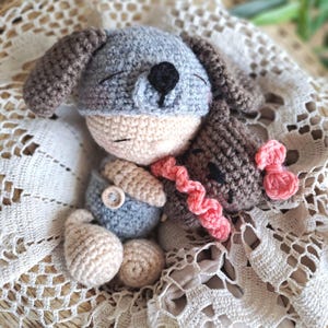 Könnte beinhalten: Eine gehäkelte Puppe mit einer grauen Hundemütze mit Schlappohren und schwarzer Nase. Die Puppe hat ein beiges Gesicht, trägt eine graue Latzhose mit einem Holzknopf und hält einen kleinen braunen gehäkelten Bären mit rosa Rüschenbesatz.