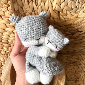 Könnte beinhalten: Zwei gehäkelte graue Katzenpuppen, eine größere und eine kleinere, umarmen sich. Die Katzen haben weiße Pfoten und rosa Akzente im Gesicht. Die Puppen sind handgefertigt und wirken weich und kuschelig.