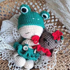 May include: A crocheted doll with a beige head and body, wearing a green frog hat with black and white eyes. The doll has a red pacifier, a green ruffled skirt with two white buttons, and holds a small brown bear with a red flower accessory.