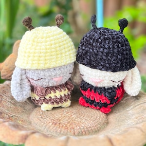 Può includere: Due peluche all'uncinetto a forma di ape con antenne. Il giocattolo a sinistra ha un cappellino giallo, testa grigia e corpo a righe marroni e gialle. Quello a destra ha un cappellino nero, testa bianca e corpo a righe rosse e nere.