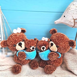 Könnte beinhalten: Zwei braune gehäkelte Teddybären, die jeweils einen kleineren braunen gehäkelten Teddybären in einer leuchtend blauen gehäkelten Tasse halten. Die größeren Bären haben weiße Schnauzen und kleine weiße Schleifen auf dem Kopf, die kleinen Bären haben weiße Schnauzen.