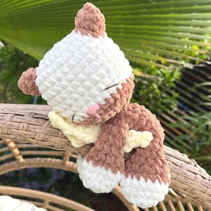 Könnte beinhalten: Ein gehäkeltes Plüschtier mit weißem Kopf, braunen Ohren und braunem Körper. Das Spielzeug hat geschlossene Augen, eine kleine rosa gestickte Nase und einen hellgelben Schal um den Hals. Die Textur ist weich und grob, mit weißen Pfoten an den Enden.