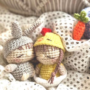 May include: Two small crochet dolls with closed eyes. One doll wears a gray acorn-shaped hat, and the other wears a yellow chick-themed hat with a red crest. A small orange crochet carrot with a green top is positioned to the right of the dolls.