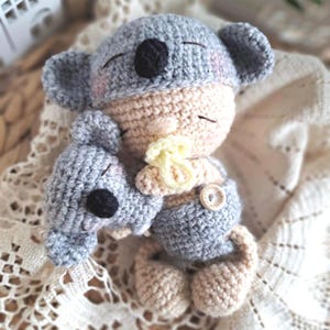 May include: A handmade crochet doll featuring a beige face with closed eyes and pink cheeks, wearing a grey koala hat with round ears and a black nose. The doll holds a smaller grey crochet koala toy and wears grey pants with a small wooden button.