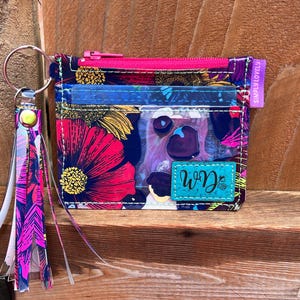 May include: A floral print card holder with a pink zipper and a matching tassel keychain. The card holder features a vibrant design with red, pink, and purple flowers on a navy blue background. The keychain has a clear plastic tassel.