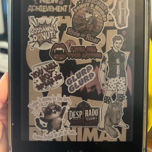 Op de afbeelding: Een zwarte Kindle e-reader met een doorzichtige beschermhoes, bedekt met stickers. Stickers bevatten tekst als "NEW ACHIEVEMENT!" en "GODDAMN! DONUT!" samen met verschillende illustraties. Het woord "kindle" is onderaan zichtbaar.