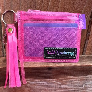 May include: A bright pink, clear wallet with a zipper closure, a key ring, and a tassel. The wallet has a purple interior and a black label that reads "Wild Dewberrys Handcrafted for You."