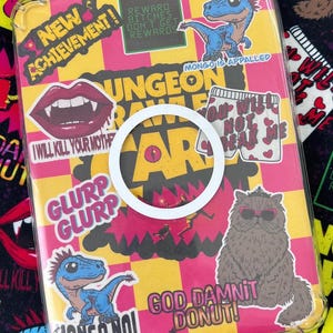 May include: A clear phone case with a colorful design featuring various stickers. The stickers include a cartoon dinosaur, a cat wearing sunglasses, and text that reads "NEW ACHIEVEMENT!" and "GOD DAMNIT DONUT!" The background is yellow and pink.