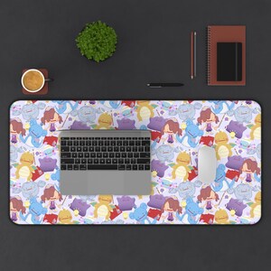 May include: A rectangular desk mat featuring a repeating pattern of cartoon characters in various colors on a light purple background. A laptop, mouse, and keyboard are on the mat. A cup of coffee, a plant, and stationery are also on the desk.